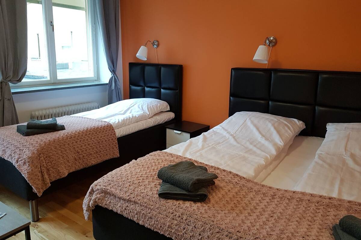 Ein Hotelzimmer mit zwei Betten, einem Fenster mit Vorhängen, einem Heizkörper und zwei Nachttischlampen.