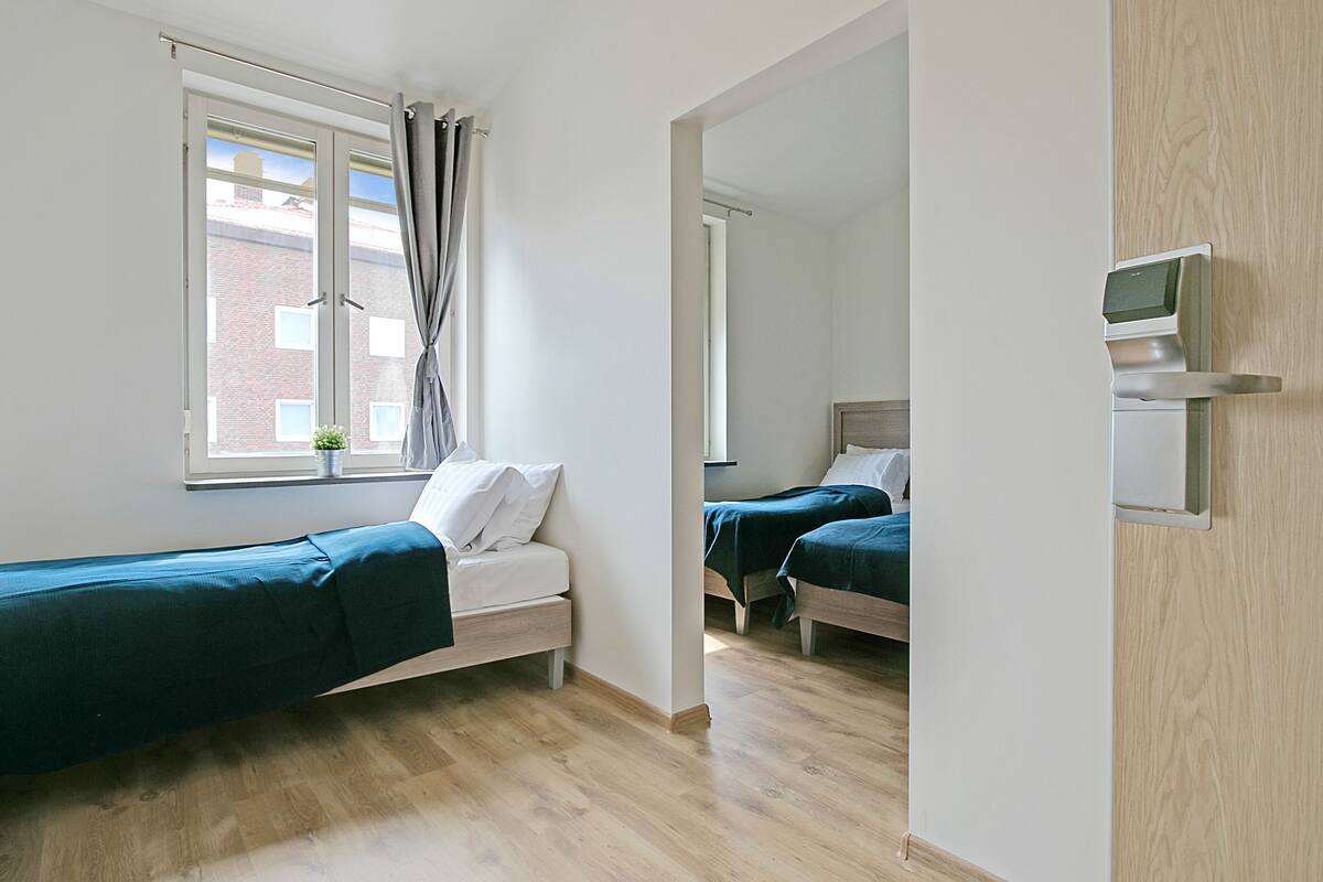 Ein Schlafzimmer mit zwei Betten, einem Fenster mit Vorhängen, einer kleinen Pflanze und einer Tür mit digitalem Schloss.