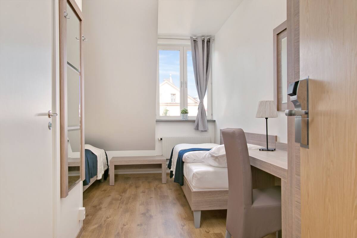 Ein kleiner, gut beleuchteter Raum mit einem Bett, einem Schreibtisch und einem Stuhl. Ein Fenster mit Vorhängen, ein Heizkörper und eine Tür sind ebenfalls vorhanden.