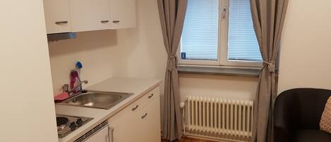 A compact kitchen with a sink, stove, and cabinets, a radiator, and a window with curtains.