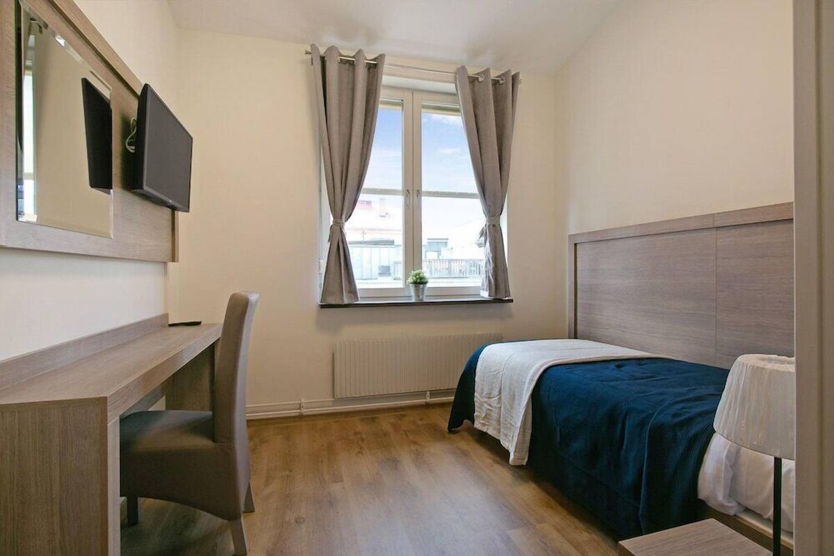 Ein Schlafzimmer mit Bett, Schreibtisch, Stuhl, einem Fenster mit Vorhängen und einer Heizung.