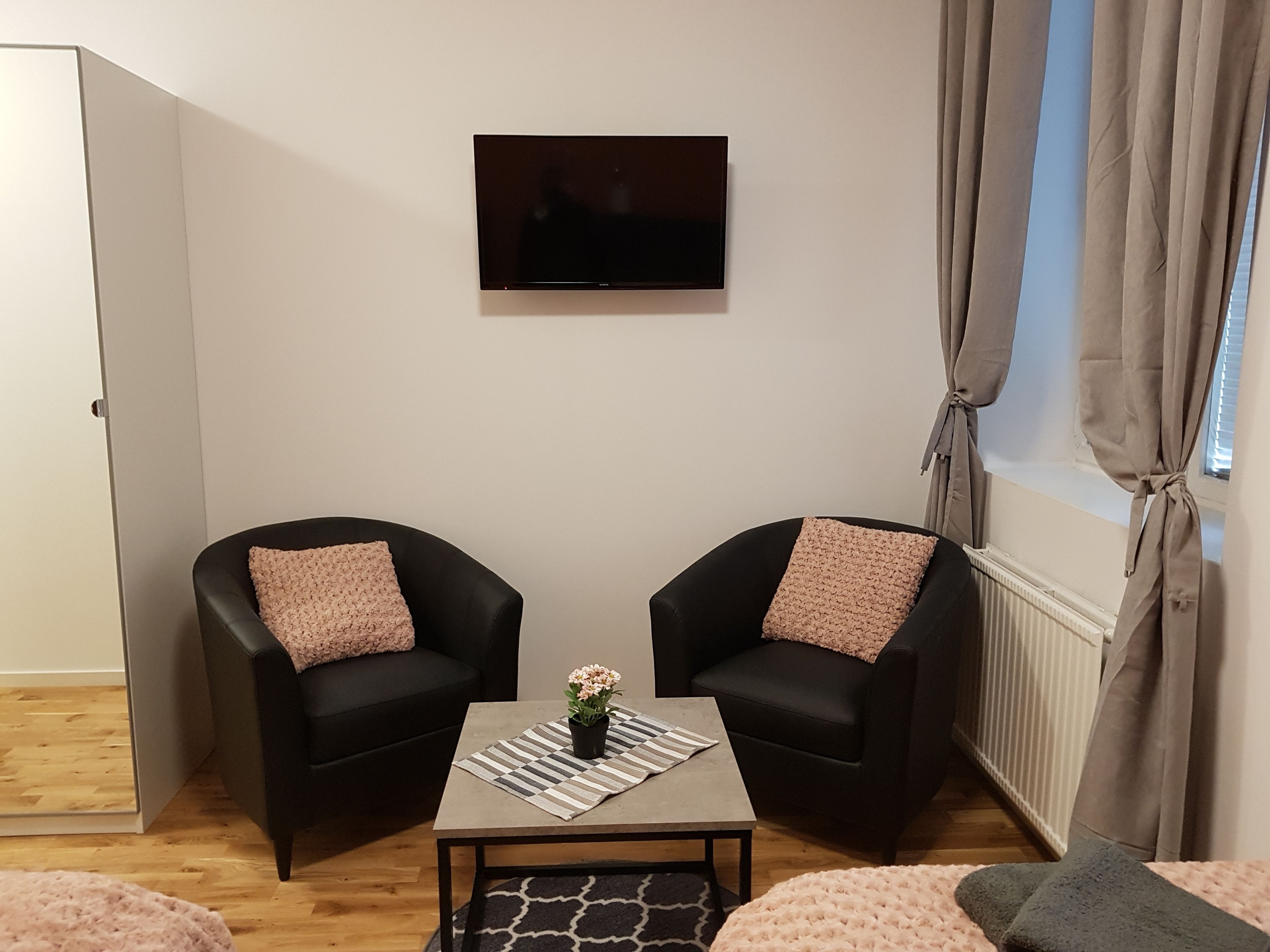 Habitació petita i moderna amb dos butaques negres, una taula amb una planta i un televisor penjat a la paret.