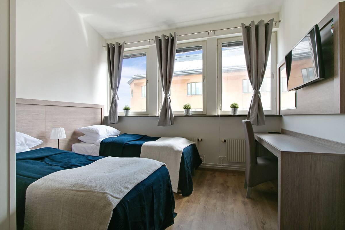 Ein Hotelzimmer mit zwei Betten, einem Schreibtisch, einem Fernseher und einem Fenster mit Vorhängen.