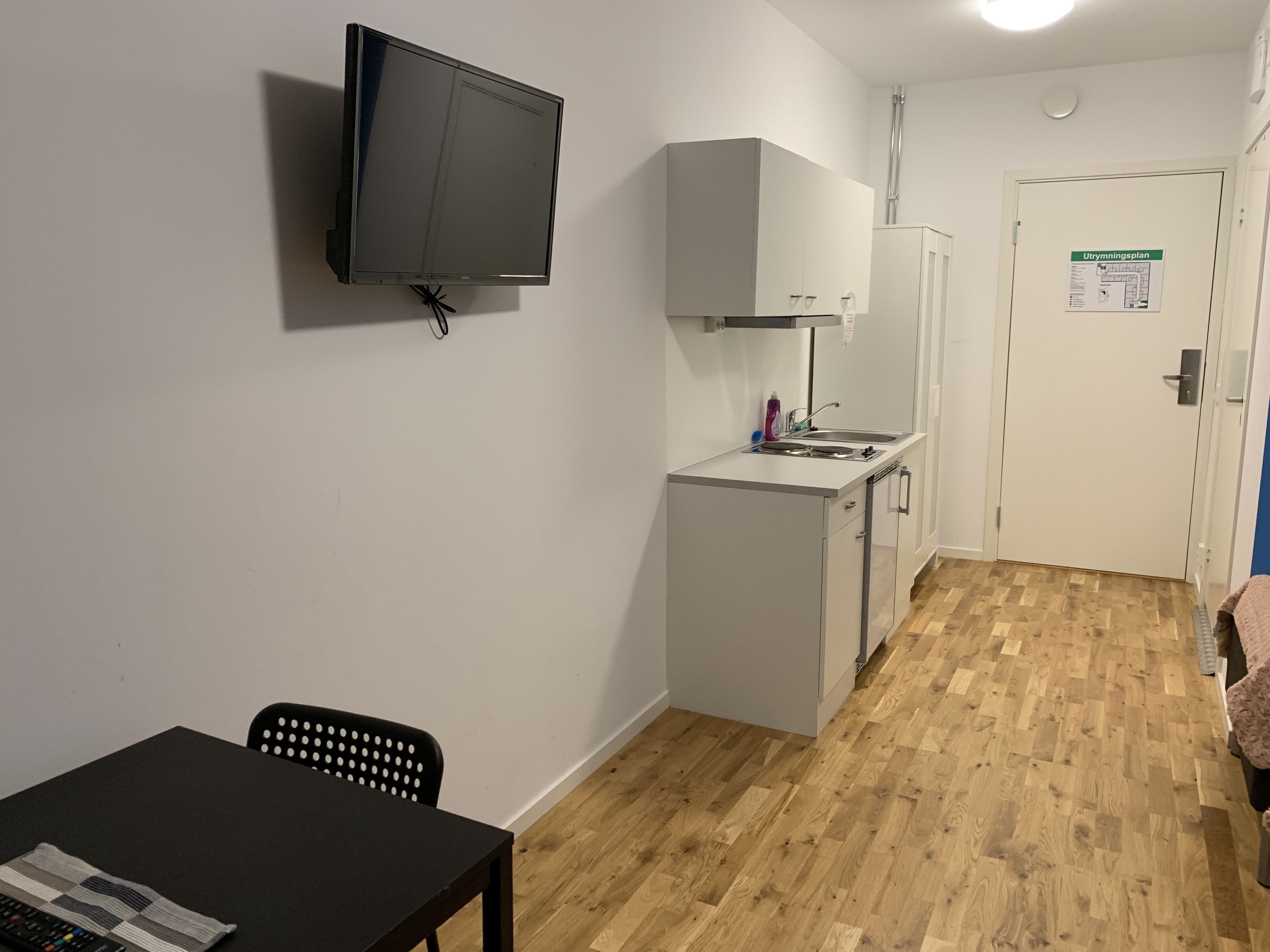 Ein kompakter Raum mit an der Wand montiertem Fernseher, einer Küchenzeile inklusive Spüle und Schränken, einem kleinen Tisch mit Stuhl und einer Tür mit Aushang.