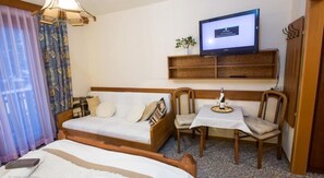 A hotel room with a bed, a sofa, a small table with a bottle and glasses, a television on a shelf, and a window with curtains.