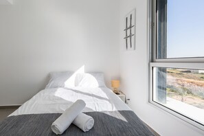 A neatly made bed with white linens and a rolled towel, a bedside lamp, and a window with a view of a desert landscape.