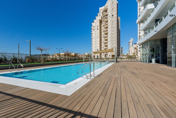 A swimming pool with a wooden deck, surrounded by modern buildings.