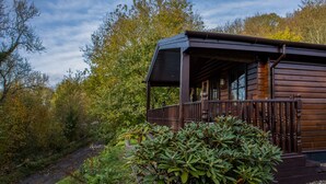 A wooden cabin with a porch, surrounded by trees and a small stream. - The Steadings (Cupar)
