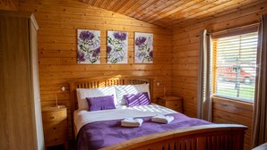 A wooden cabin bedroom with a bed, nightstands, and a window with blinds. - The Steadings (Cupar)