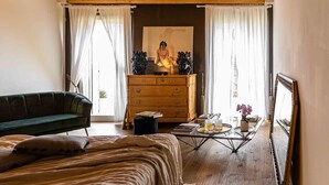 Une pièce chaleureuse avec un canapé vert, une commode en bois, un lit recouvert d’une couverture beige, une table basse avec des livres et un vase de fleurs, et un tableau accroché au mur.