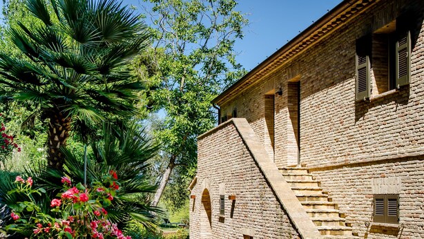 Un bâtiment en briques avec un escalier menant à un étage supérieur, entouré d’une végétation luxuriante et de plantes en fleurs.