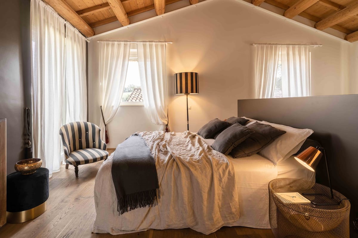 Une chambre à coucher avec un lit, une chaise, une lampe et une fenêtre avec des rideaux.