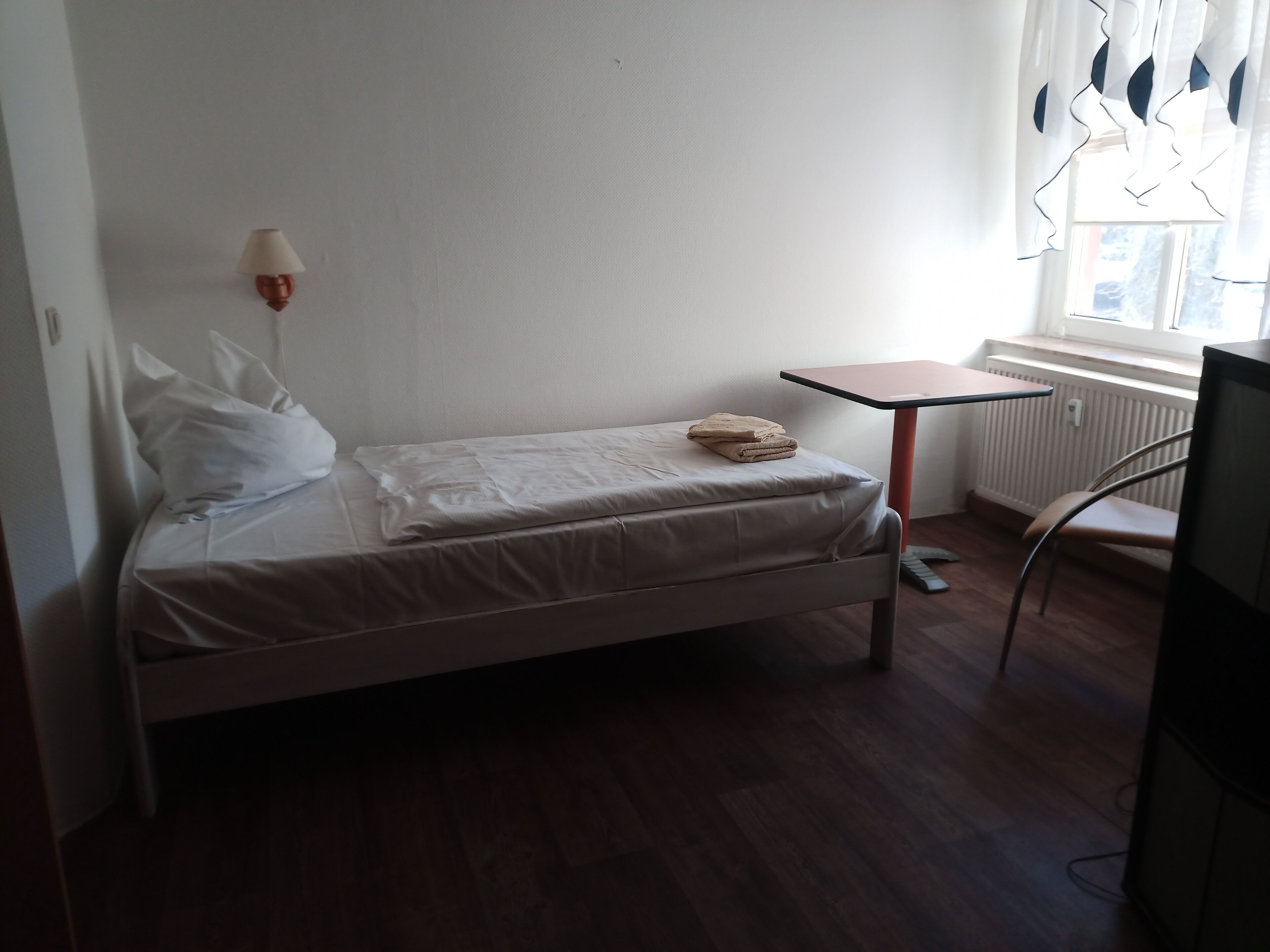 A single bed with a white mattress and pillows, a small table with a lamp, and a chair.