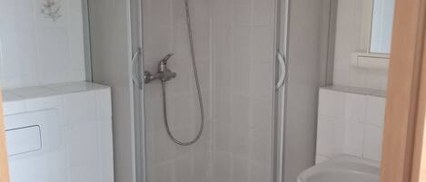Ducha en esquina con puerta de vidrio, inodoro blanco y lavabo con espejo superior.