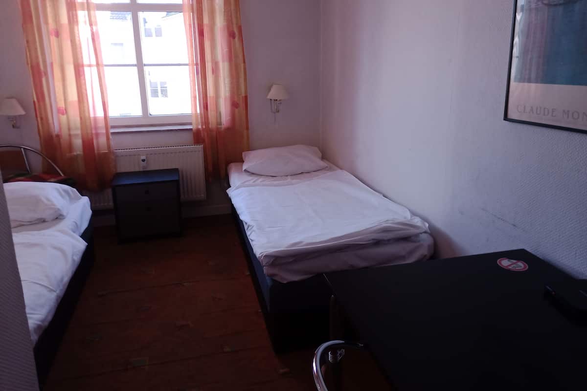 A small, single-room hotel room with two beds, a desk, a chair, a radiator, and a window with curtains.