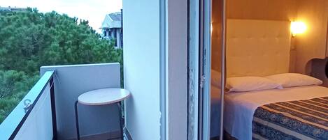 A balcony with a small table and chairs, a sliding glass door to a bedroom with a bed and a nightstand, and a view of greenery and buildings in the background.