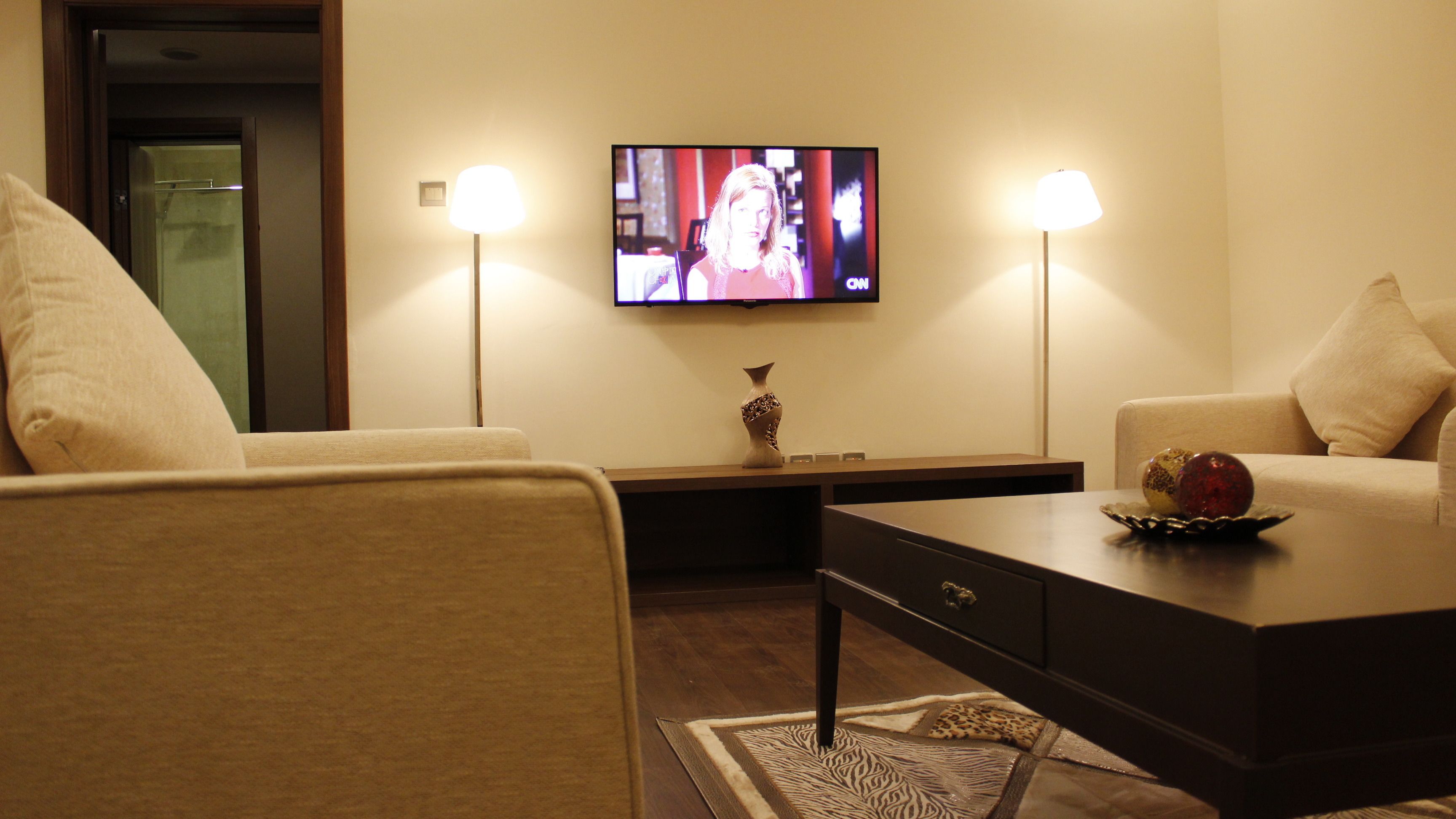 A living room with two beige sofas, a dark coffee table, a vase, and a flat-screen TV mounted on the wall.