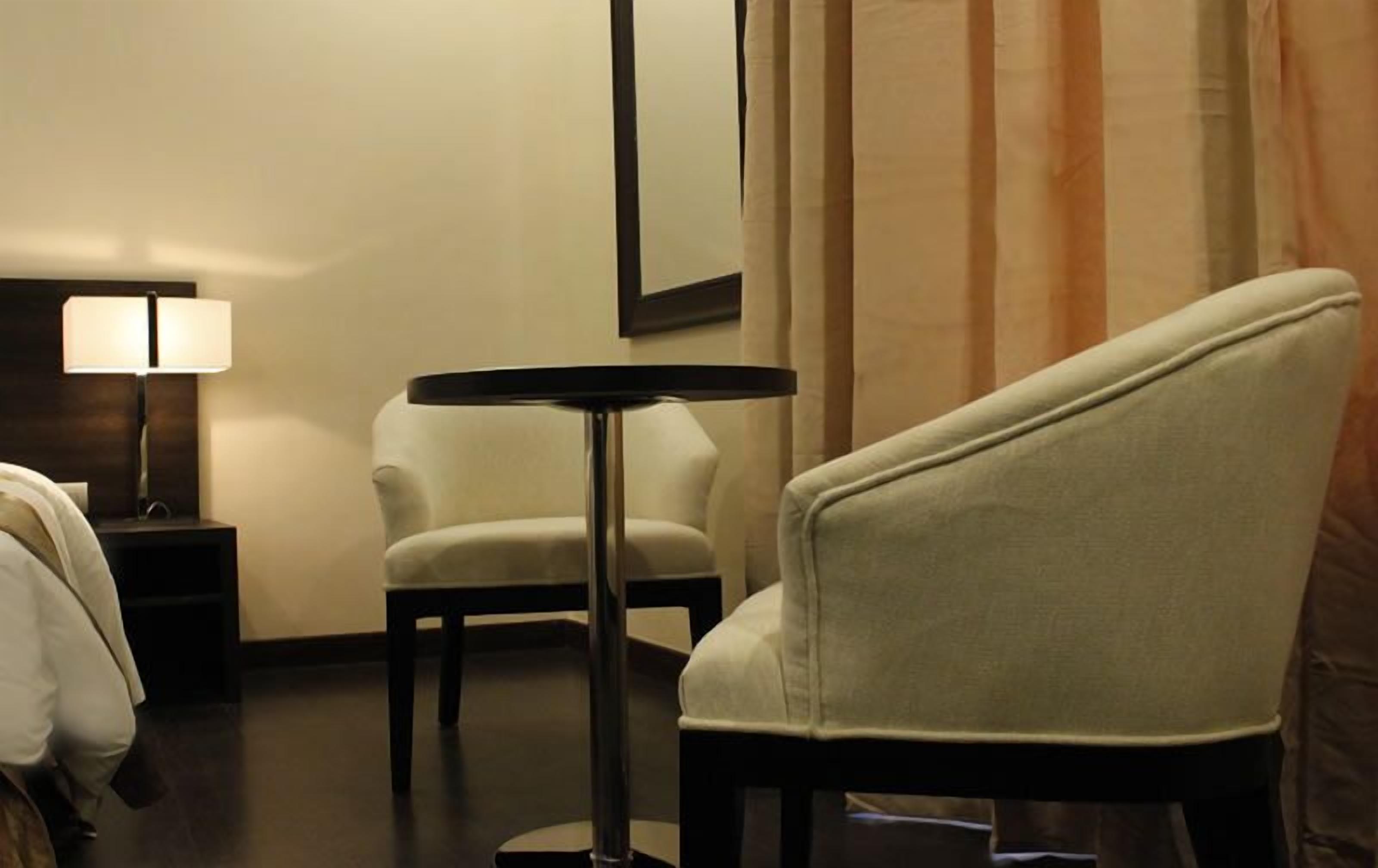 A hotel room with a single white armchair, a small round table, a lamp, and a bed with white linens.