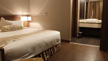 A hotel room with a neatly made bed, a bedside lamp, a small bedside table, and a view into the bathroom through an open door.