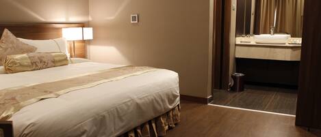 A hotel room with a neatly made bed, a bedside lamp, a small bedside table, and a view into the bathroom through an open door.