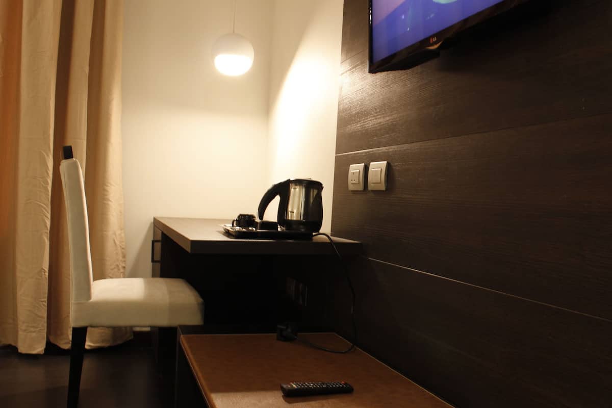 A hotel room with a wooden desk, a chair, a remote control, a kettle, and a television mounted on the wall.