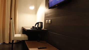 A hotel room with a wooden desk, a chair, a remote control, a kettle, and a television mounted on the wall.