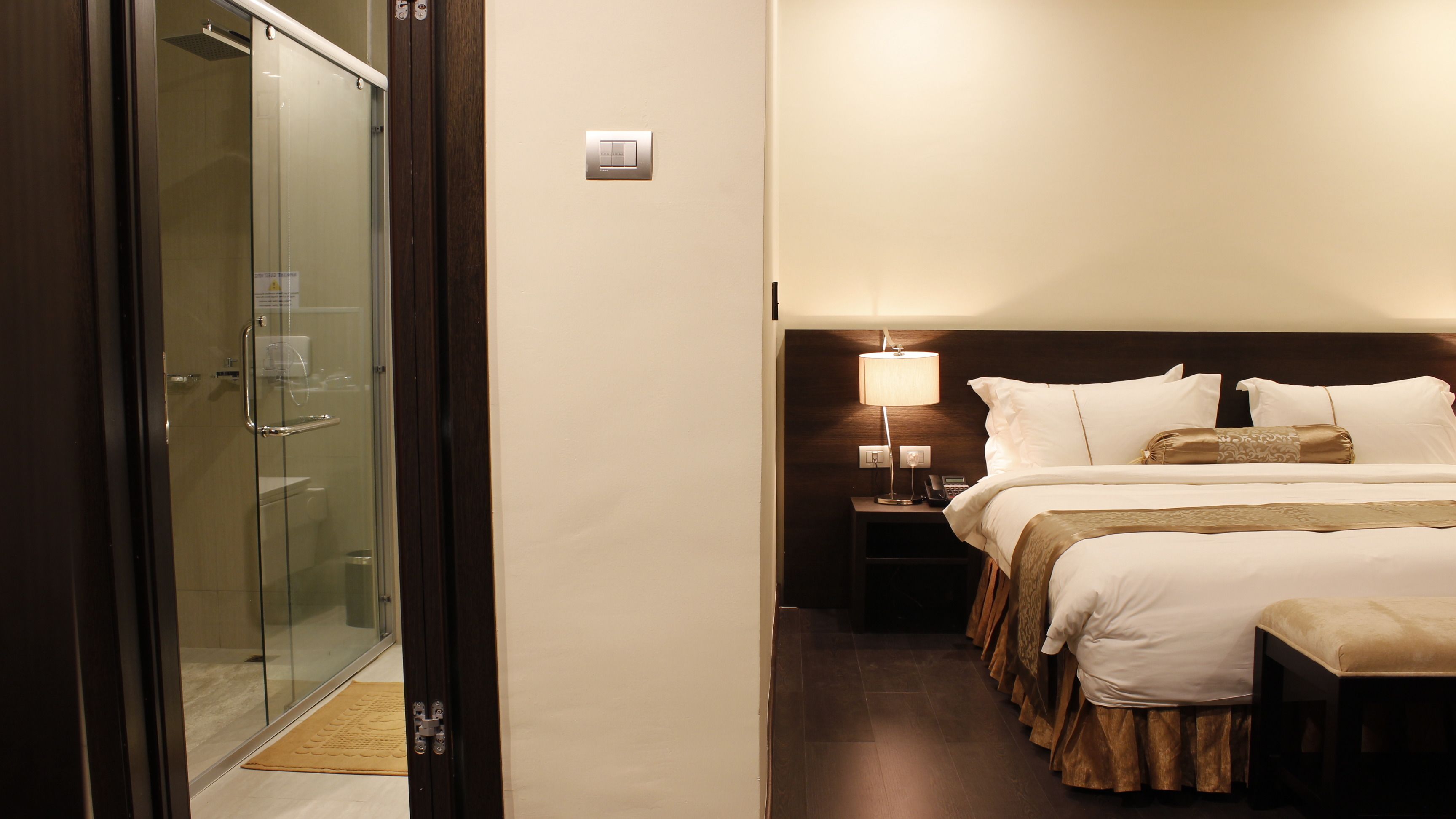 Habitación de hotel con puerta de vidrio que da a la zona de ducha, una mesita de noche con lámpara y una cama bien tendida con cabecera.