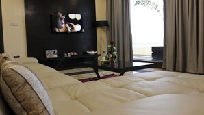 Uma sala de estar moderna com um amplo sofá bege, uma televisão de tela plana na parede, uma mesinha lateral com objetos decorativos e vista para o exterior através de uma janela com cortinas.