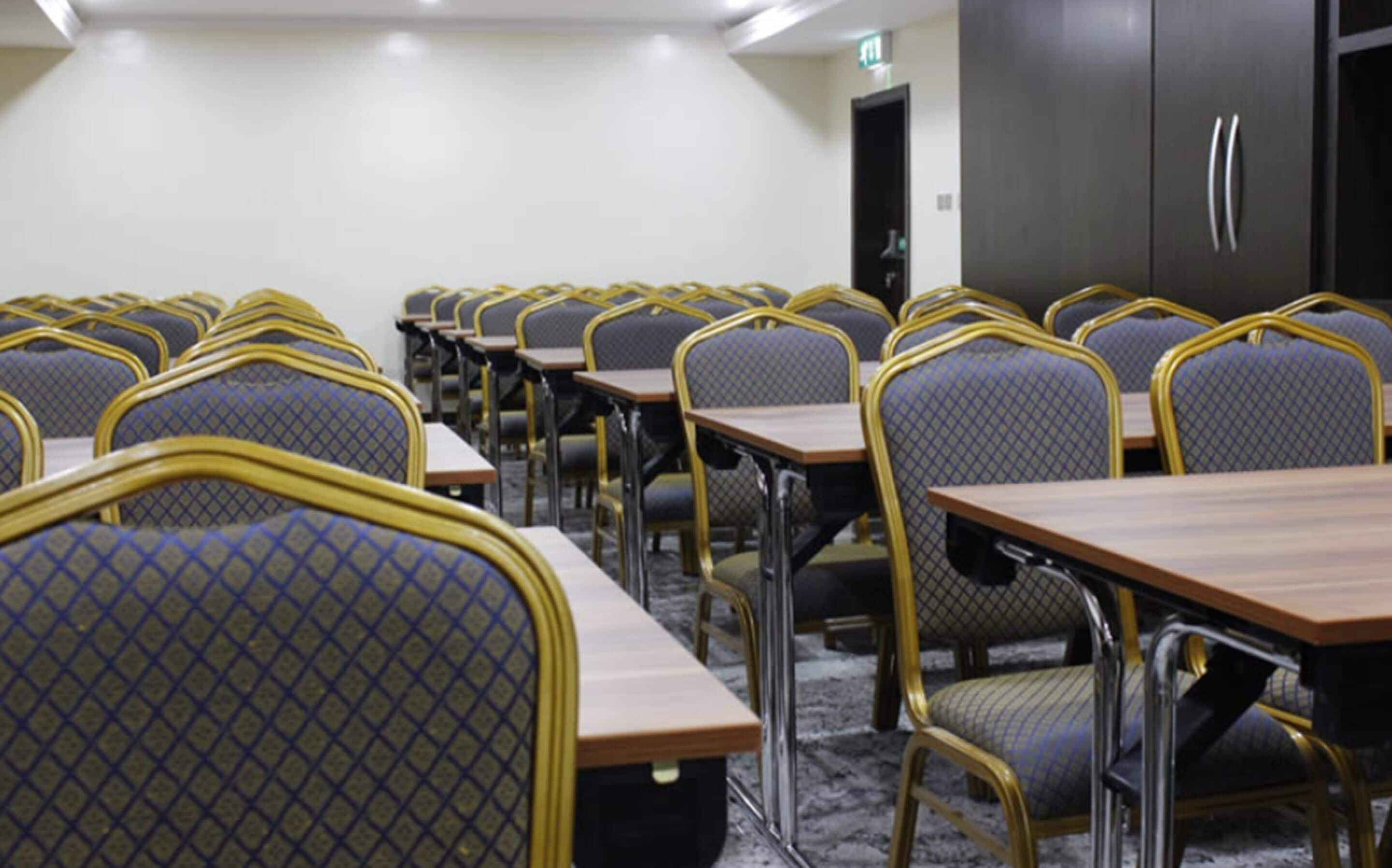 Una sala de conferències amb files de taules i cadires, equipada amb una pantalla de projector i una porta.