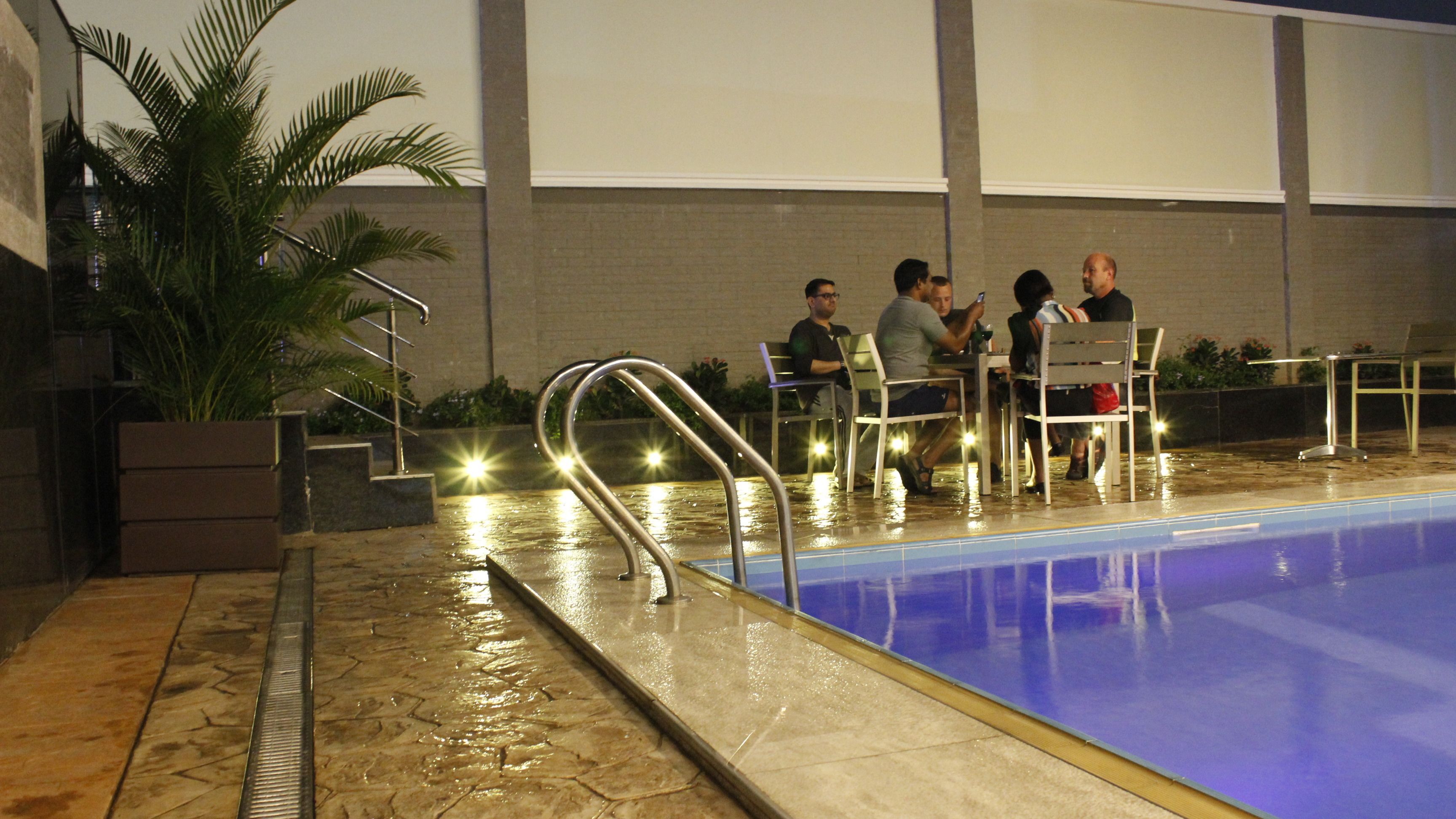 A swimming pool with a tiled deck, metal railings, and a few people seated around a table.