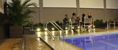 A swimming pool with a tiled deck, metal railings, and a few people seated around a table.