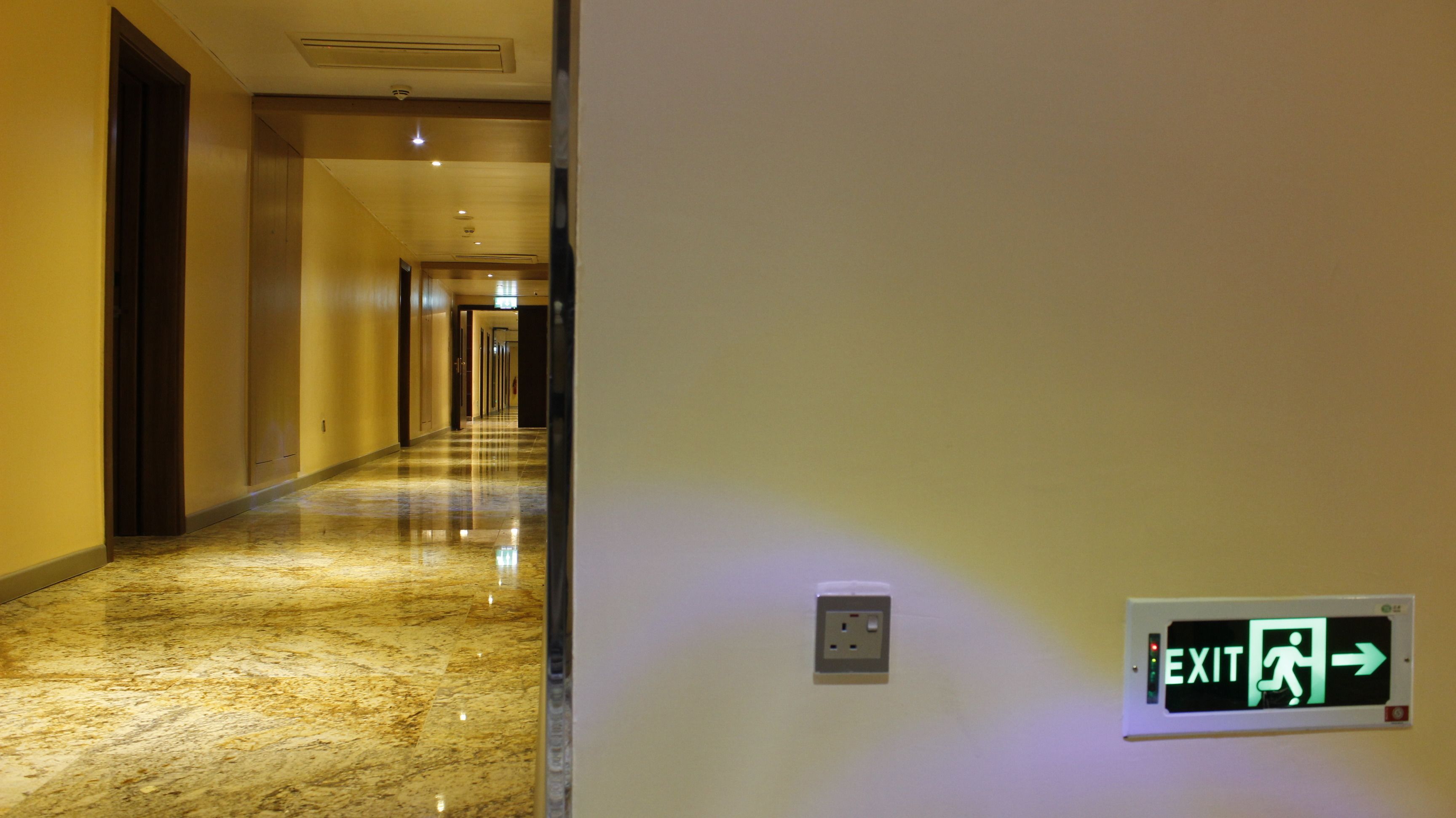 Un pasillo de hotel con piso de mármol pulido, un letrero de salida en la pared y una puerta a la izquierda.