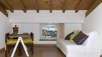 Een kamer met een witte bank, een eettafel met stoelen, een raam met uitzicht op een stadsgezicht en een houten plafond.