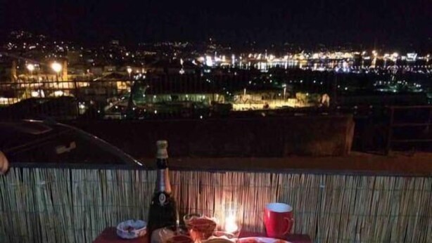 A table with a bottle, glasses, and plates of food, overlooking a cityscape at night. - La Finestra sul Golfo Affittacamere (La Spezia)