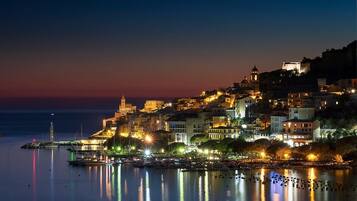 Un pueblo costero al atardecer con edificios iluminados, una torre destacada y un mar tranquilo que refleja las luces.