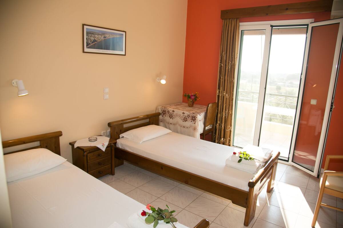 Ein Hotelzimmer mit zwei Einzelbetten, einem kleinen Tisch mit Blumenvase, einem Fenster mit Vorhängen und Blick ins Freie.