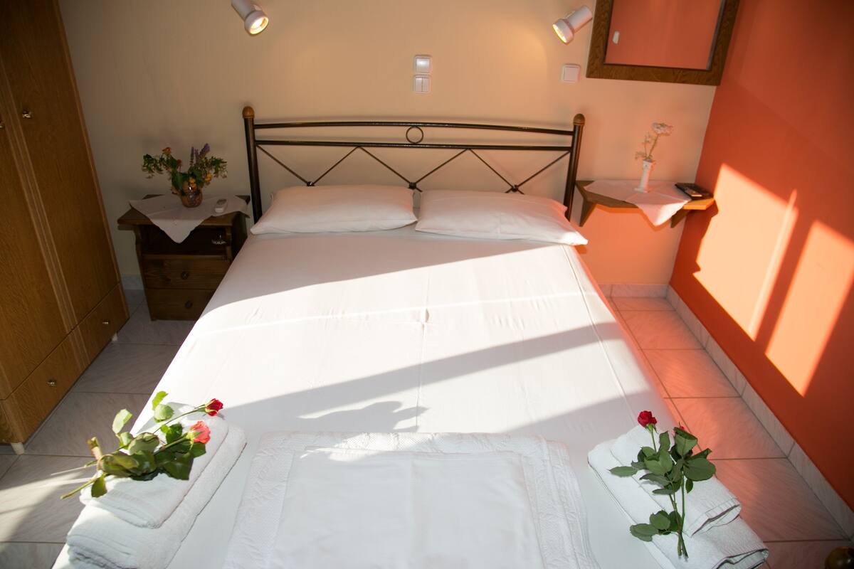 Ein ordentlich bezogenes Bett mit weißen Leinen und Kissen, ein Nachttisch mit Blumenvase und auf der rechten Seite des Bettes ein kleiner Tisch ebenfalls mit Blumenvase.