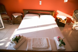 A neatly made bed with white linens and two towels with roses, a wooden nightstand, a chair, and a small table with a lamp. - Almyros Studios & Apartments (Kefalonia)