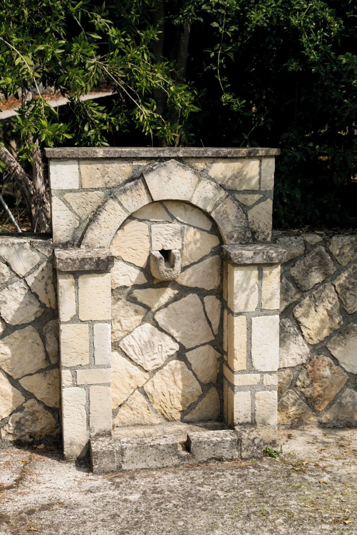 Estructura de piedra con una entrada arqueada y una pequeña abertura, rodeada de un muro de piedra y follaje.