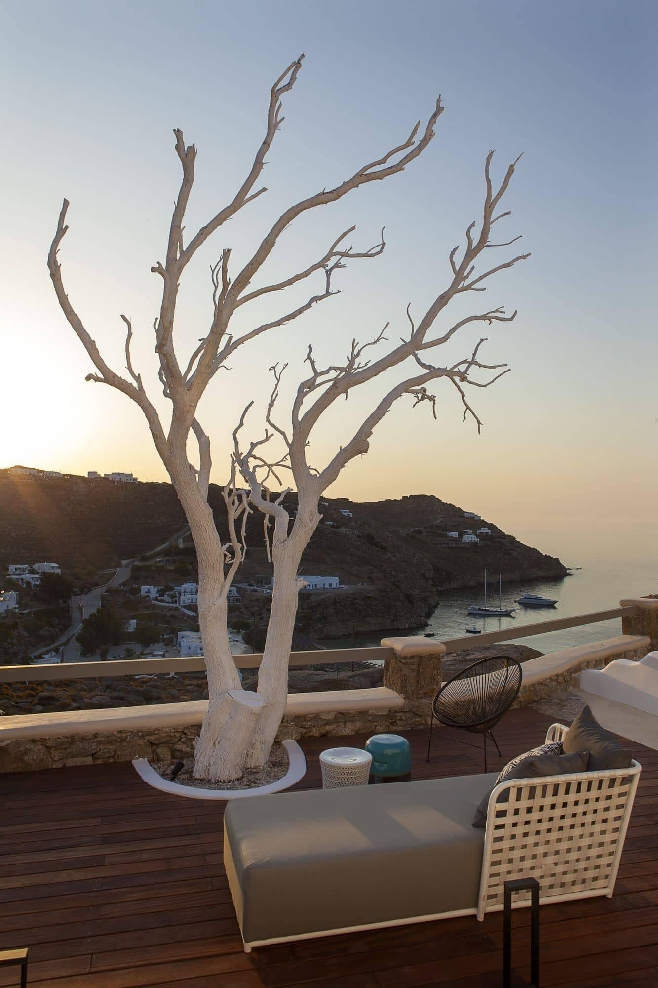 A terrace with a white tree, a white sofa, and a chair, overlooking a coastal landscape with buildings and boats.