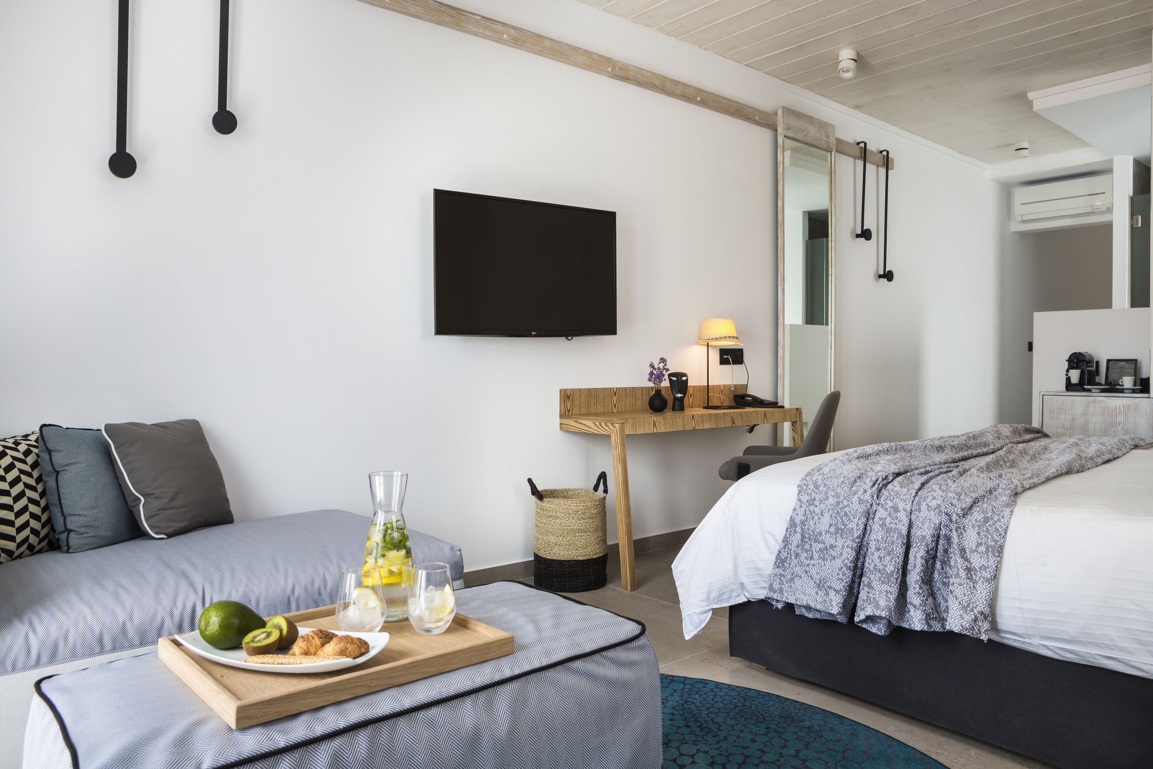 Una habitación de hotel moderna con cama, escritorio con lámpara, televisor y bandeja de desayuno con fruta y pasteles.