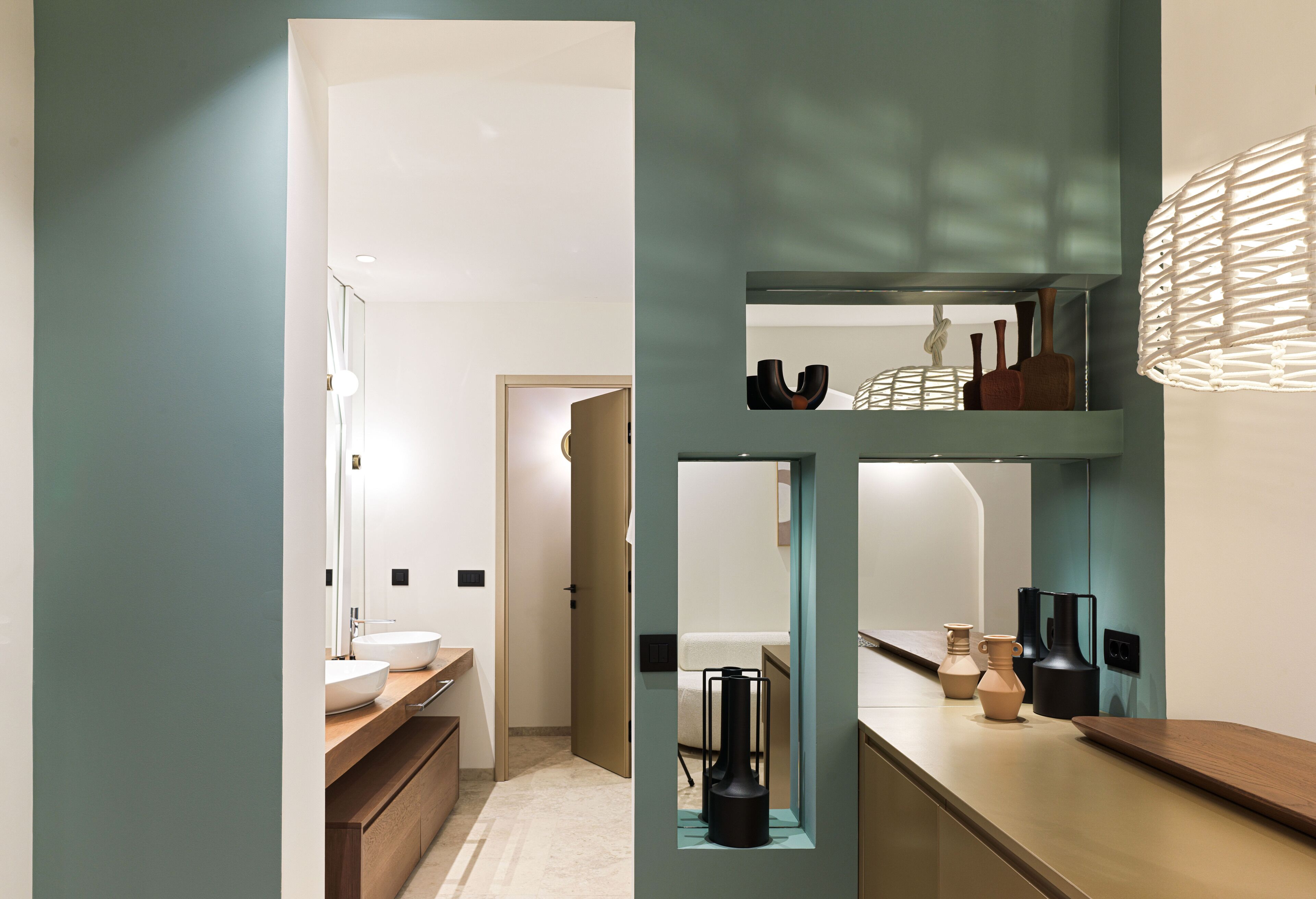 Un baño moderno con un espejo grande, un lavamanos y un tocador. Hay una puerta que conduce a otra habitación.