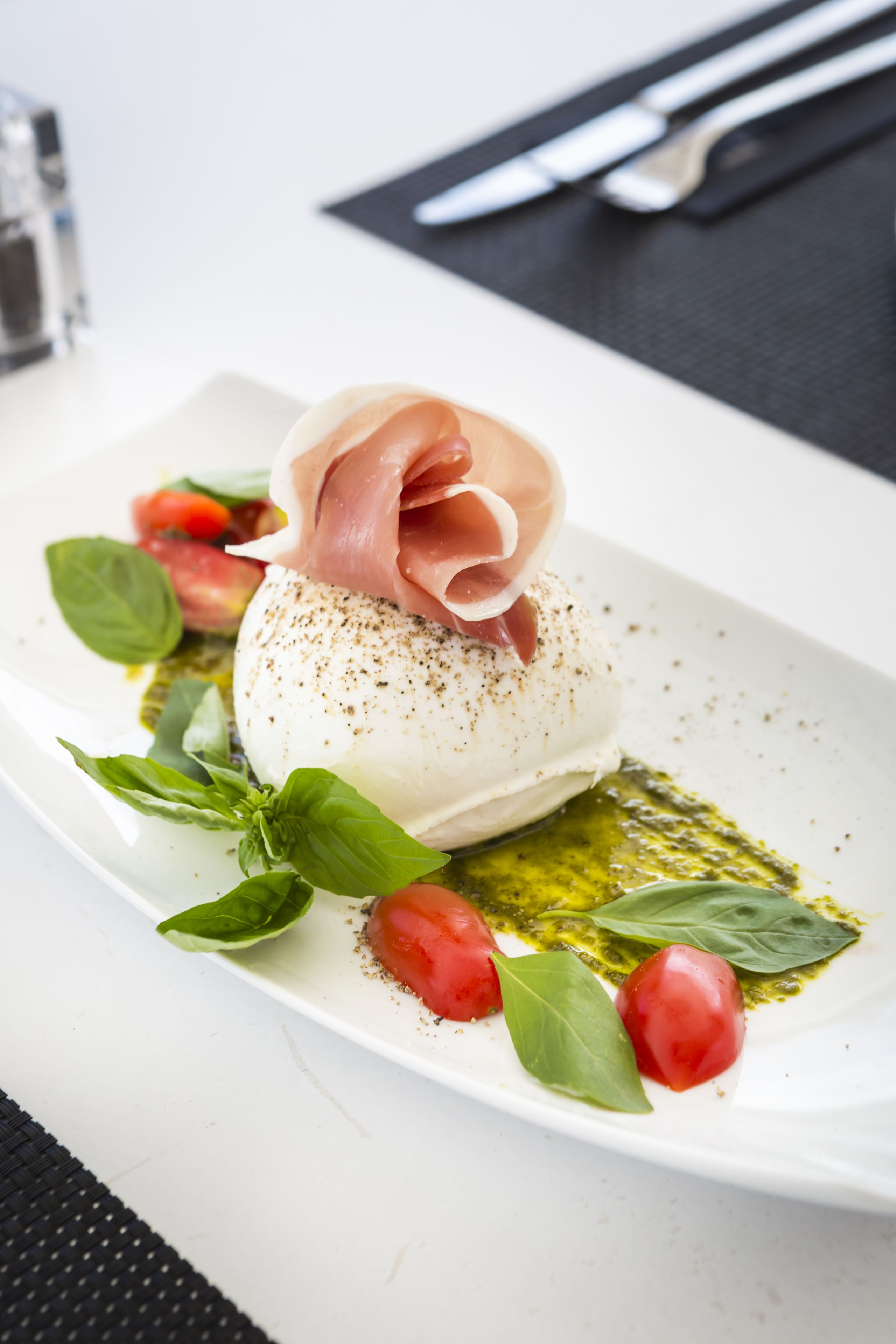 Un plato blanco con queso blanco redondo cubierto de prosciutto, adornado con hojas de albahaca y tomates cherry, y un chorrito de salsa verde.