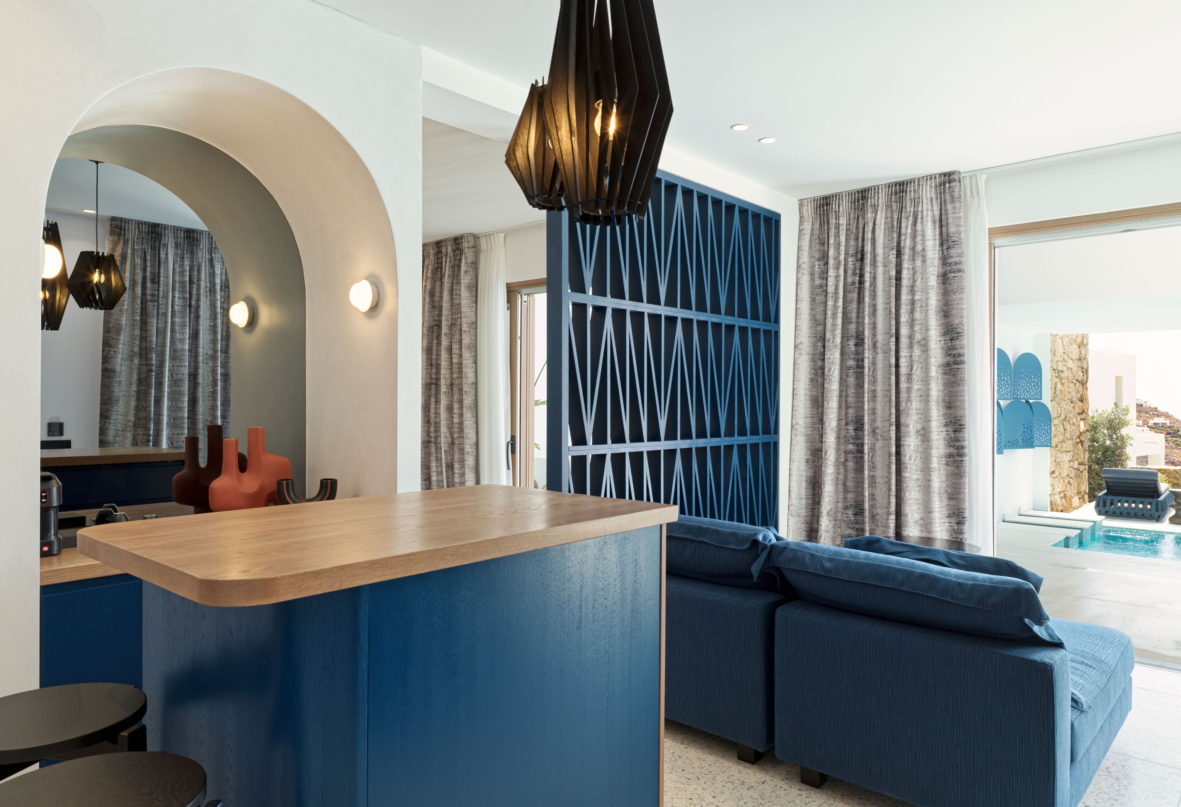 A modern living room with a blue accent wall, a wooden coffee table, a blue sofa, and a large window with a view of a pool.