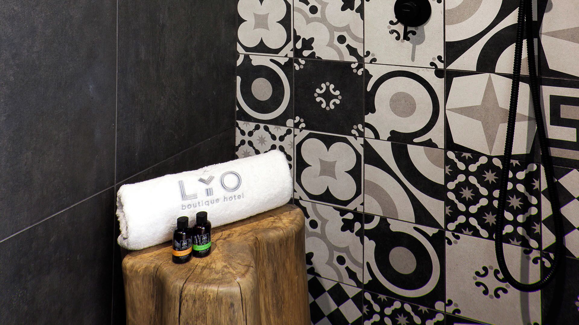 A bathroom with a wooden stool, a white towel with "LYO boutique hotel" text, and two bottles on the stool.