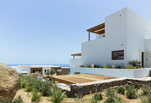 A modern white building with a wooden deck, stone walls, and a view of the sea.