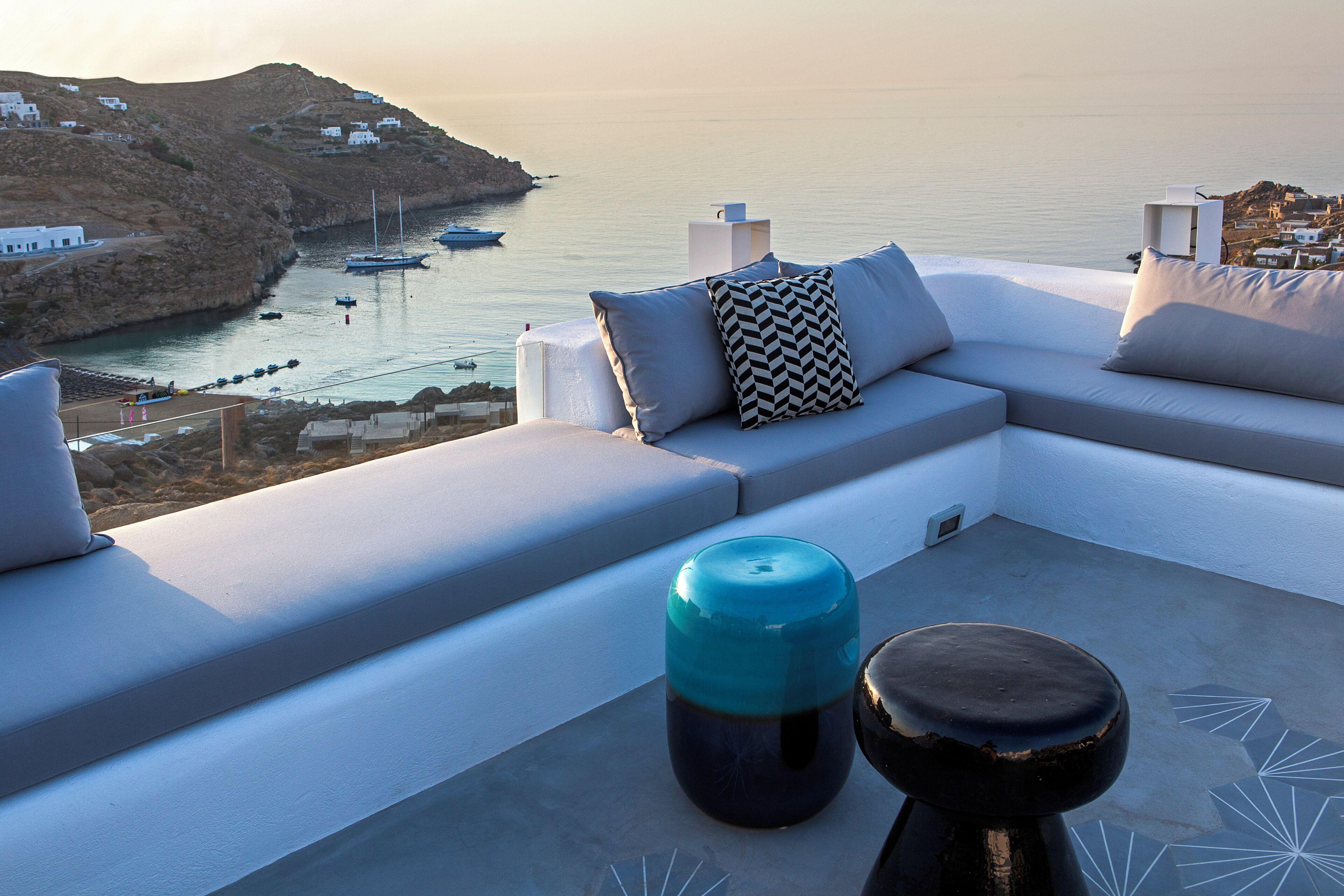 A modern outdoor seating area with a sofa and cushions, overlooking a coastal view with boats and buildings.