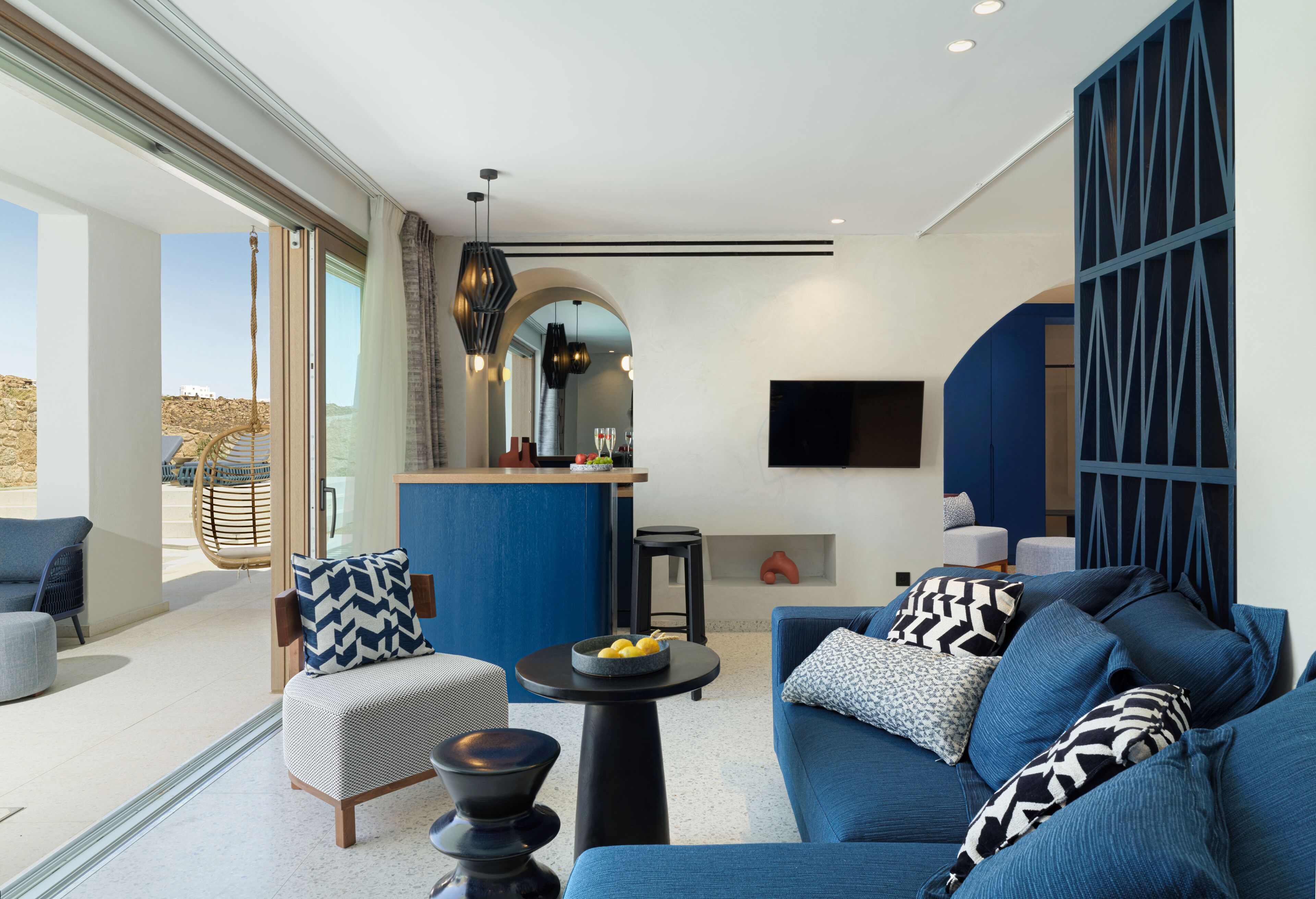 A modern living room with a blue sofa, a round coffee table, and a wicker chair. There is a kitchen area with a bar and a TV mounted on the wall.