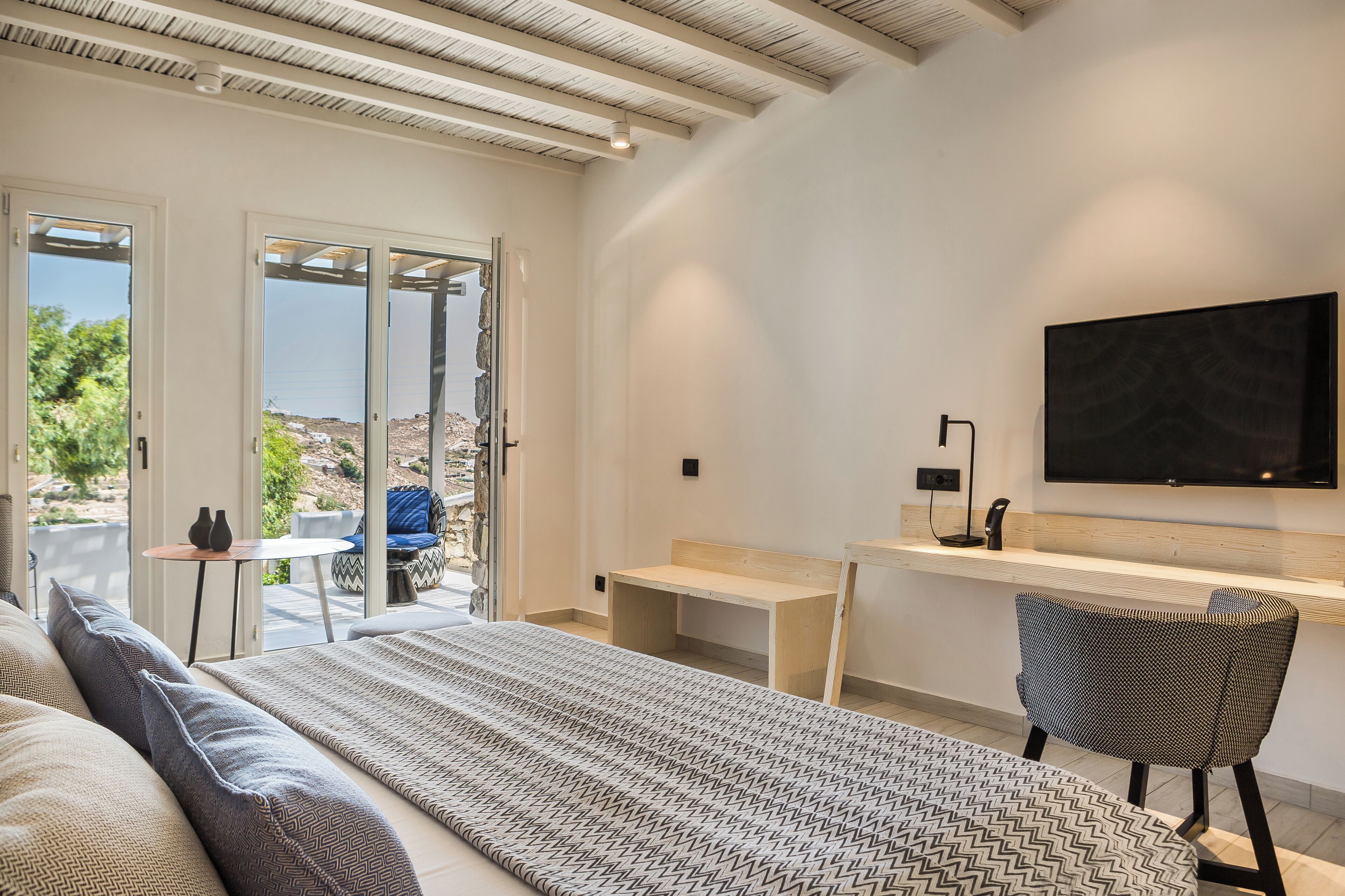 A modern bedroom with a bed, a desk, and a chair. There is a TV mounted on the wall and a window with a view of greenery.