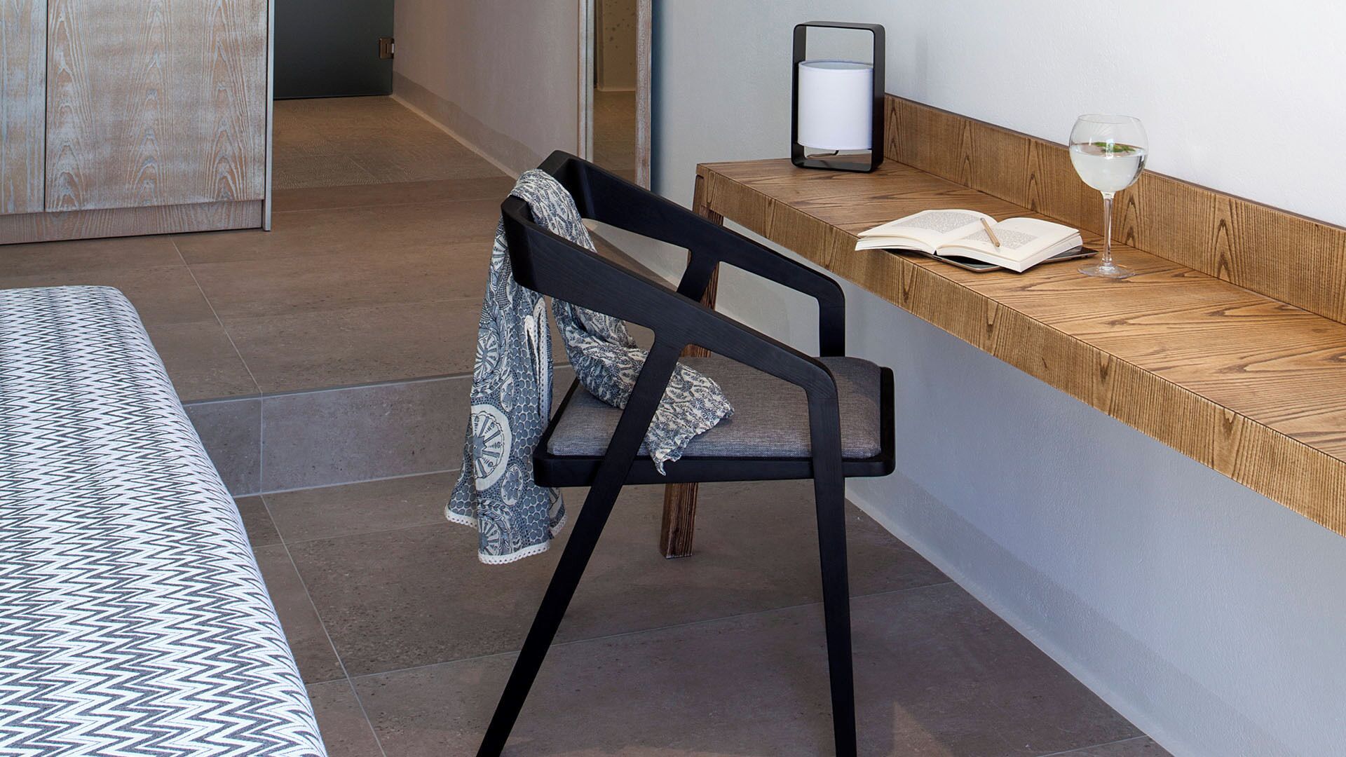 A modern hotel room with a bed, a wooden bench, a chair, and a bedside table with a lamp and a glass of water.